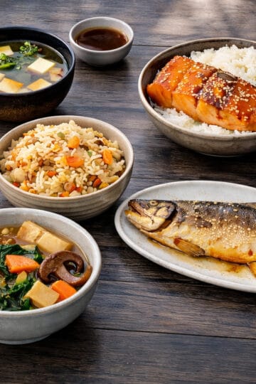 Essential Japanese cuisine dishes featuring miso soup, takikomi gohan, teriyaki salmon, salt-grilled fish, and simmered vegetables on a dark oak butcher block.