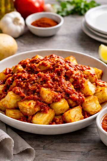 Patatas bravas with crispy potato chunks and traditional smoked paprika bravas sauce in a shallow bowl on grey-stained oak butcher block, Spanish tapas style