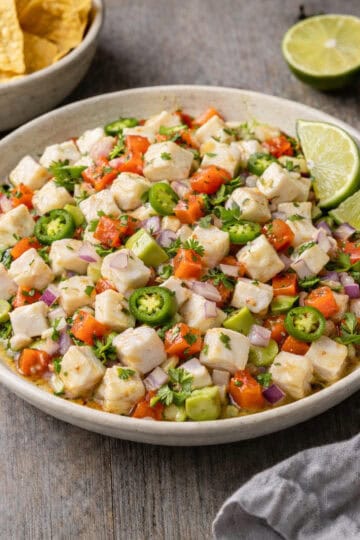 Citrus-marinated white fish ceviche with red onion, jalapeño, avocado, and lime in a shallow bowl on grey-stained oak butcher block, daylight food photography