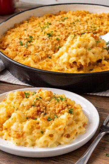 Mac and cheese in an enameled cast iron pan with a scoop removed, served on a plate on a butcher-block kitchen counter.