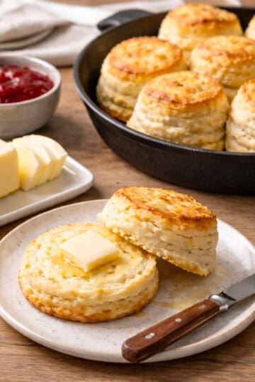 Buttermilk biscuits baked in a cast iron skillet on a light grey-stained oak butcher block counter, with one biscuit split open on a plate, sliced butter on a butter dish, a small bowl of jam, and a spreading knife arranged in a tight, rustic composition.