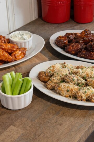 Assorted chicken wings in buffalo, BBQ, and parmesan garlic styles on a rustic wooden counter with celery and carrot sticks.