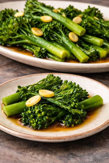 Garlic broccolini served on a platter with a small plate in the foreground, photographed on a grey-stained wood kitchen counter.