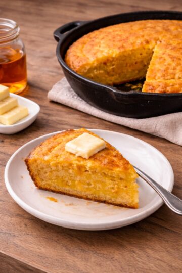 Old-fashioned Southern cornbread baked in a cast iron skillet with a slice served on a plate with butter and honey on a wood countertop.