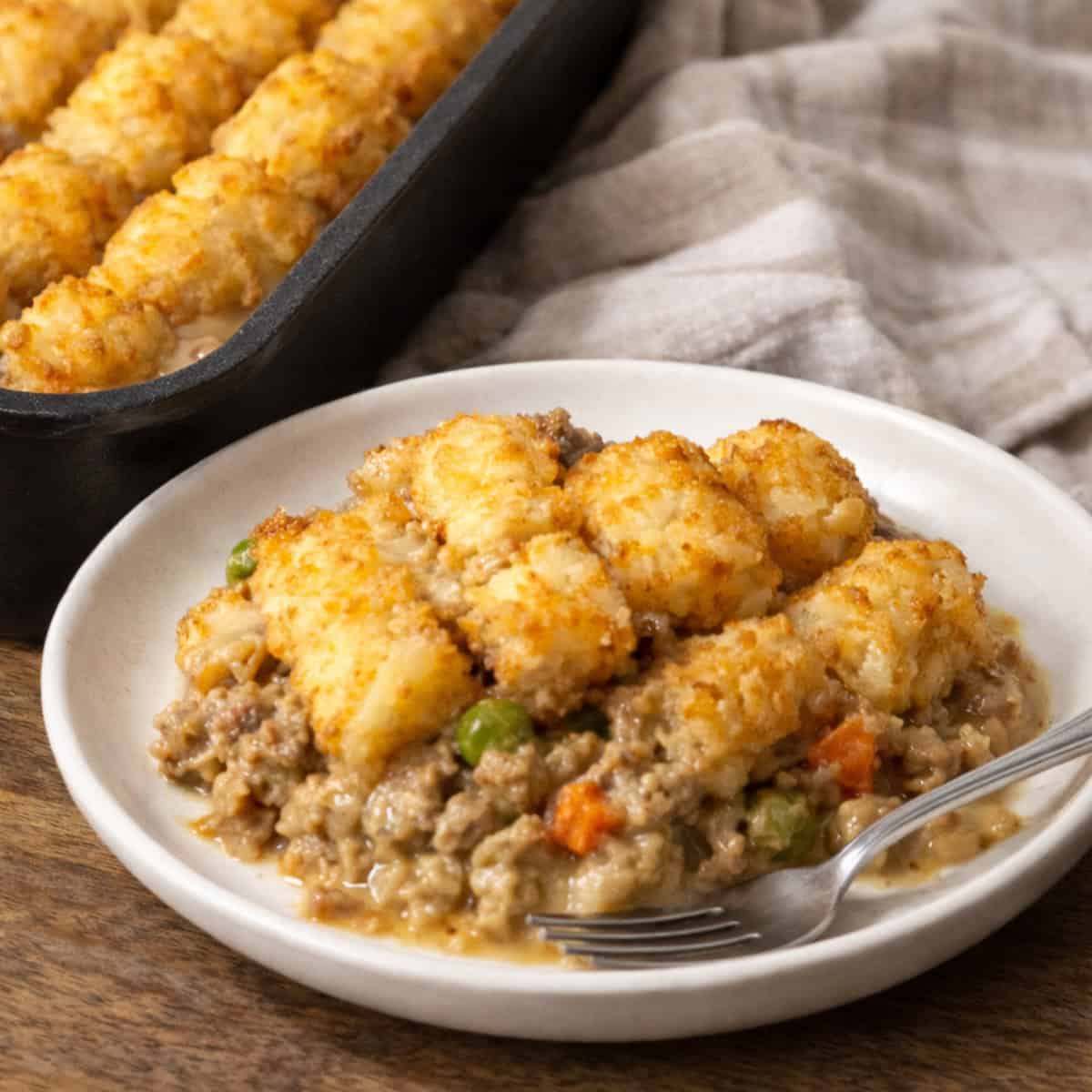 Close-up of Midwest tater tot hotdish with crispy golden tater tots over creamy ground beef and vegetables, served on a white plate on a grey-stained oak butcher block counter.