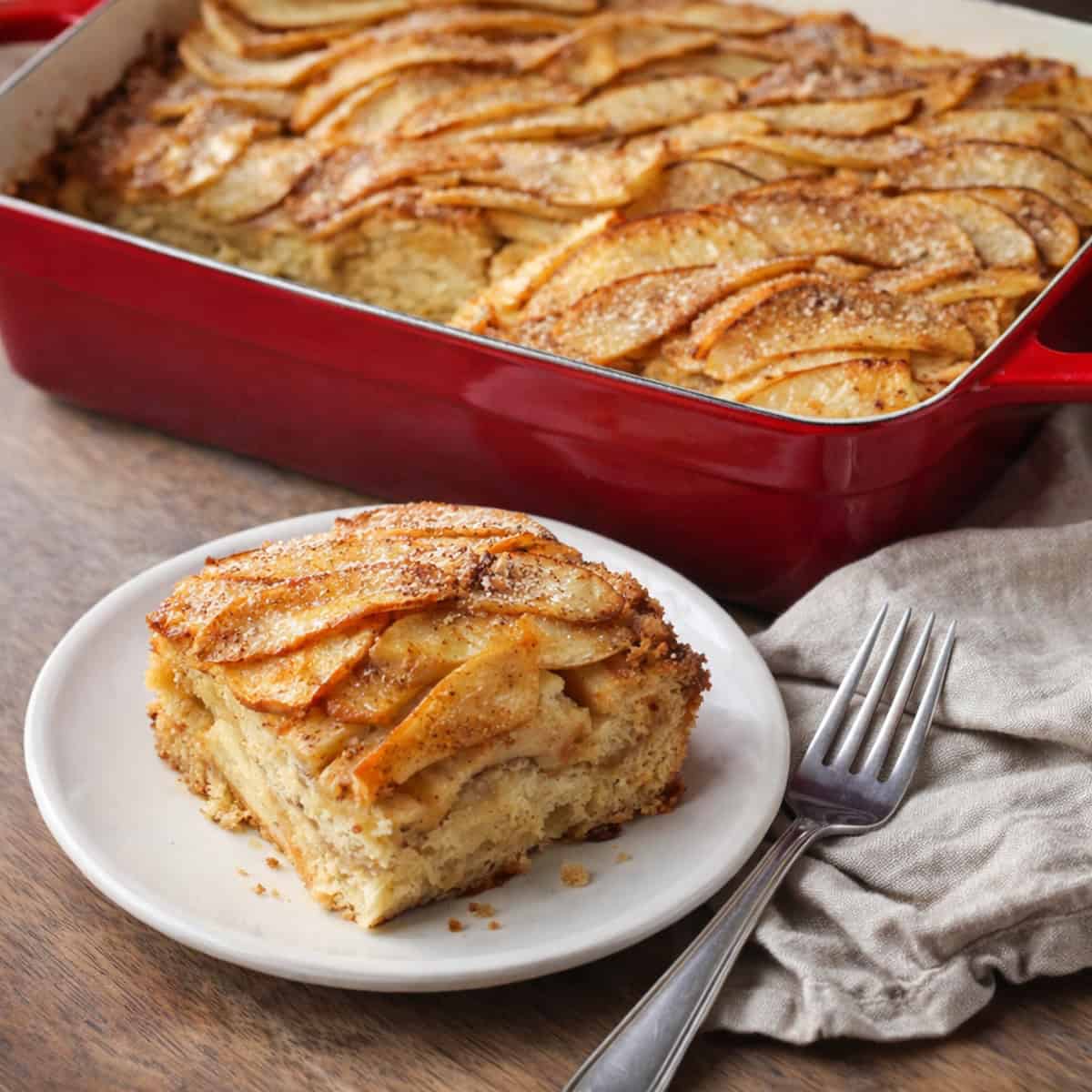 Apple French toast casserole in a red ceramic baking dish with thinly sliced apples on top, a corner slice removed, and a matching piece served on a white plate with a fork and rumpled napkin on a wood countertop.