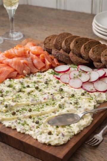 Smoked salmon butter board with thick spread of shallot and caper butter topped with fresh dill, served with cocktail-size brown bread slices, radishes, and a delicate fork on a wood countertop with champagne glasses in the background.